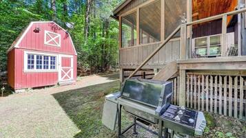 Une grange rouge et une maison en bois dotée d’une terrasse, entourées d’une cour verdoyante et d’arbres.