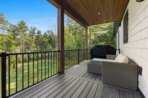 Une terrasse extérieure couverte, équipée d’un barbecue, de meubles en osier et offrant une vue sur une zone boisée.
