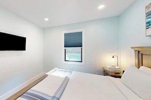 A hotel room with a bed, a bedside table with a lamp, a window with blinds, and a mounted TV. - New 5-BR Home with Movie Theater, Bar, Game Room (Lake George)