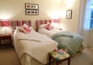 Ein Hotelzimmer mit zwei Betten, jeweils mit Kissen und einem Nachttisch mit Lampe. An der Wand hängen gerahmte Bilder.