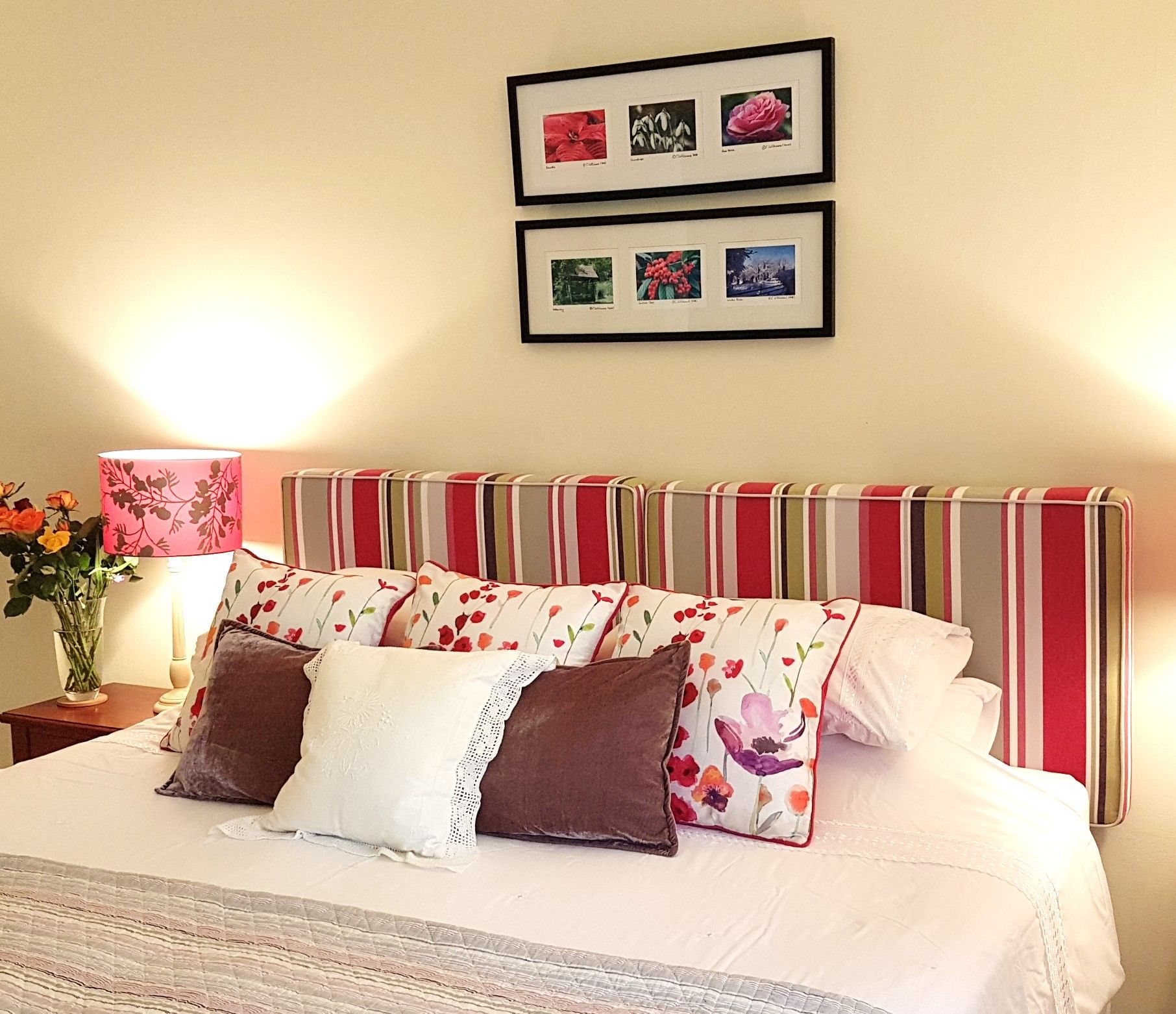 Een net opgemaakt bed met bloemenkussens, een gestreept hoofdeinde en ingelijste foto’s aan de muur.