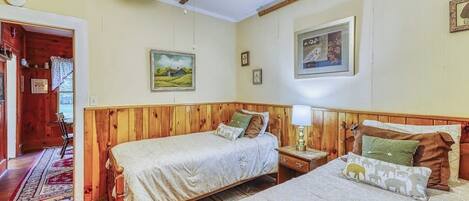 A bedroom with two beds, wooden headboards, a nightstand, a lamp, and a painting on the wall.