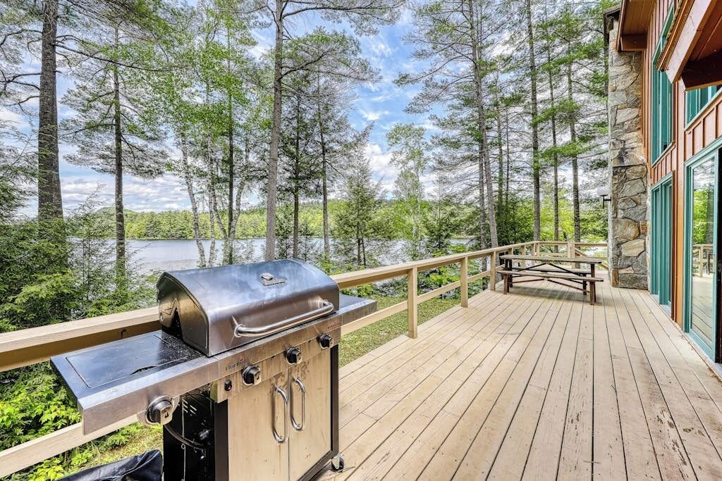 A wooden deck with a grill, a picnic table, and a view of a forest and lake.