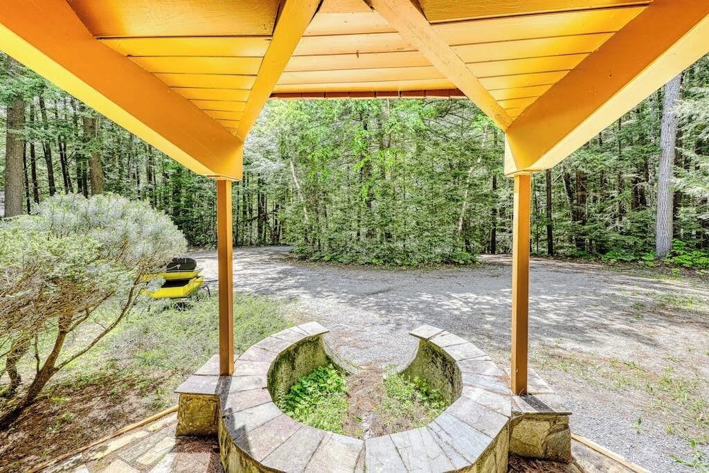 A covered outdoor area with a circular stone structure, surrounded by a forested environment.