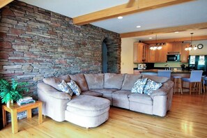 A living room with a stone wall, a sectional sofa, a small table with books, and a kitchen in the background.