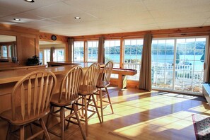 A wooden dining area with a large table and chairs, large windows with a view of a lake, and a wooden ceiling.
