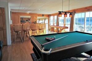 A pool table with four wooden chairs, a bar area, and large windows with a view.