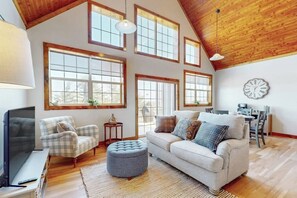 A spacious living room with a high ceiling, large windows, a television, a sofa, a chair, and a dining area. - 1BR vineyard stay with firepit, grill, & EVC (Keuka Park)