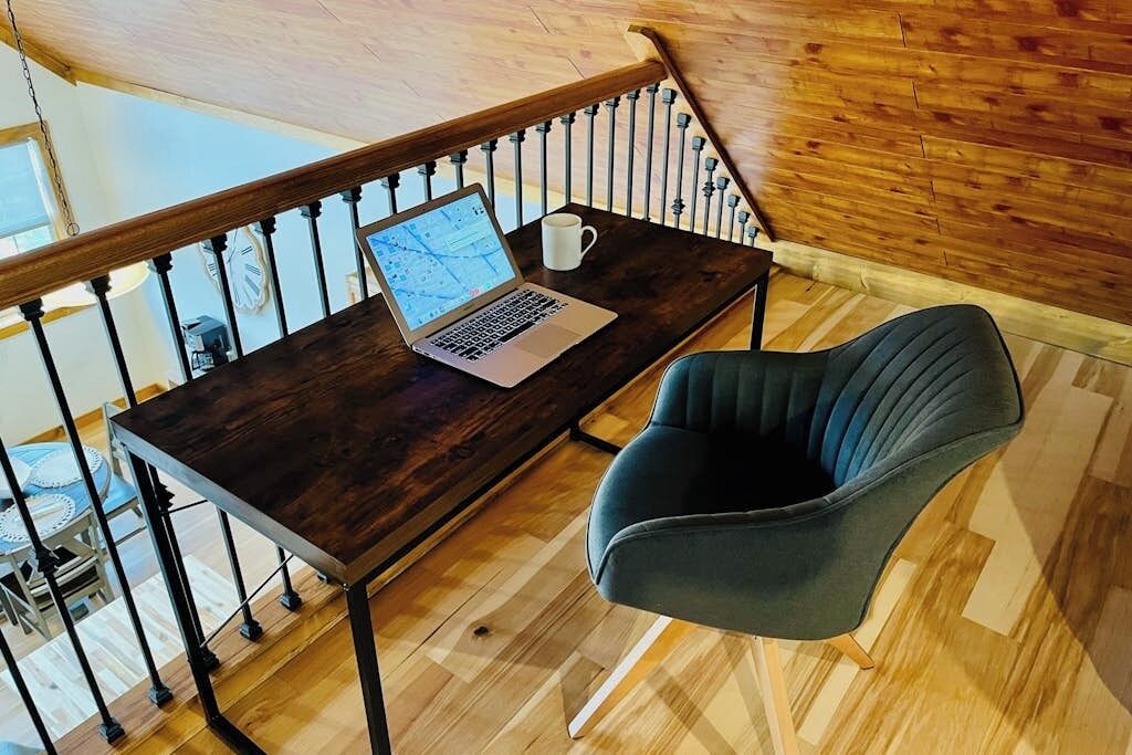 Un ordinateur portable est posé sur une table en bois sombre, à côté se trouve une tasse blanche, une chaise bleue et une rambarde en bois.