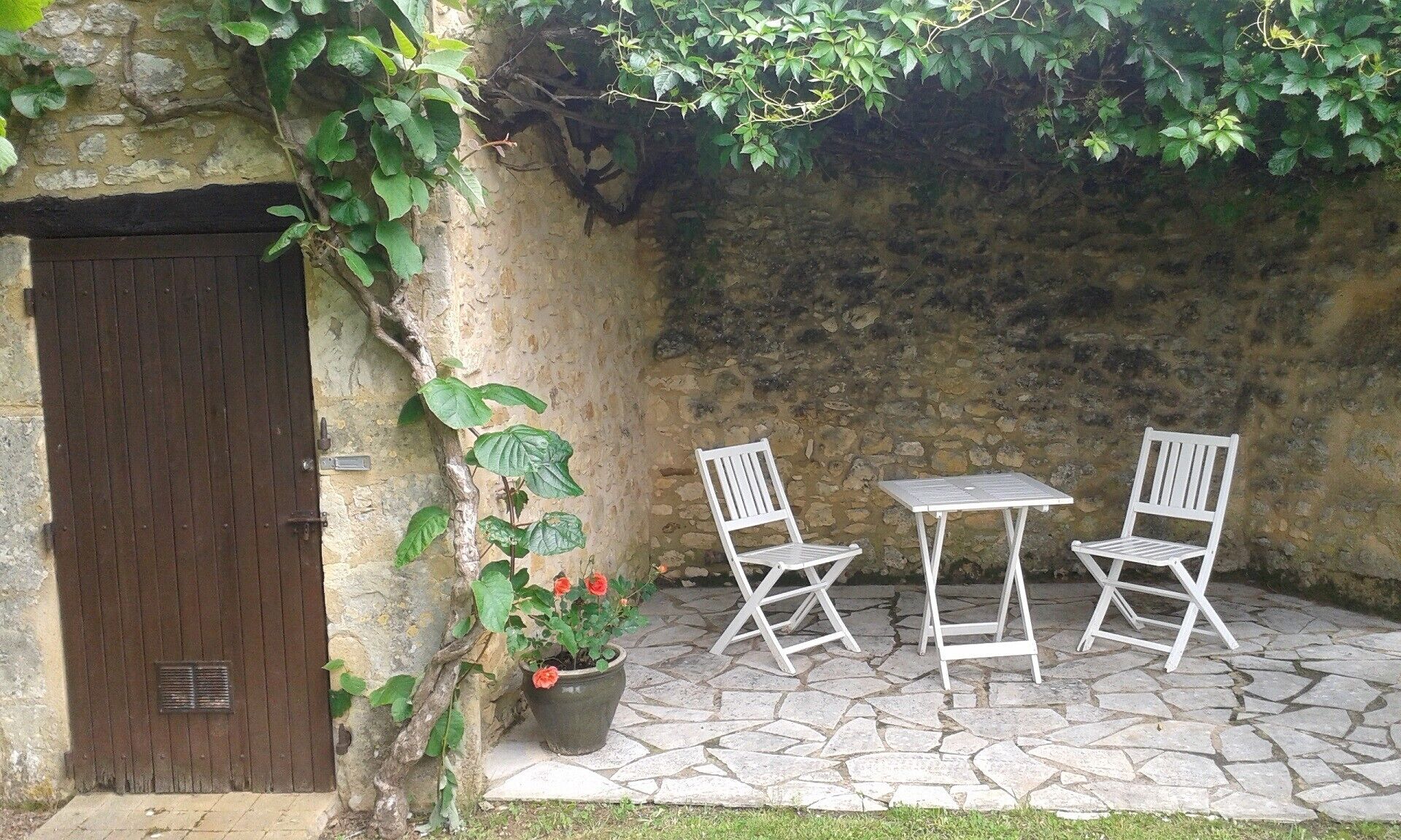 A stone patio with two white chairs and a table, a potted plant with red flowers, and a wooden door.