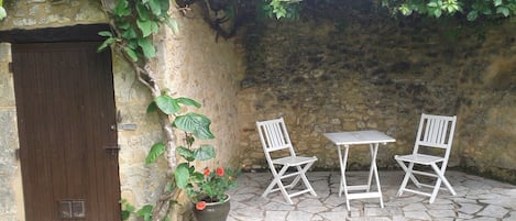 Un patio de piedra con dos sillas blancas y una mesa, una planta en maceta con flores rojas y una puerta de madera.