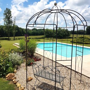 Ein Metall-Pavillon mit Sitzbank, Schwimmbecken und einem angelegten Garten mit verschiedenen Pflanzen und Steinen.