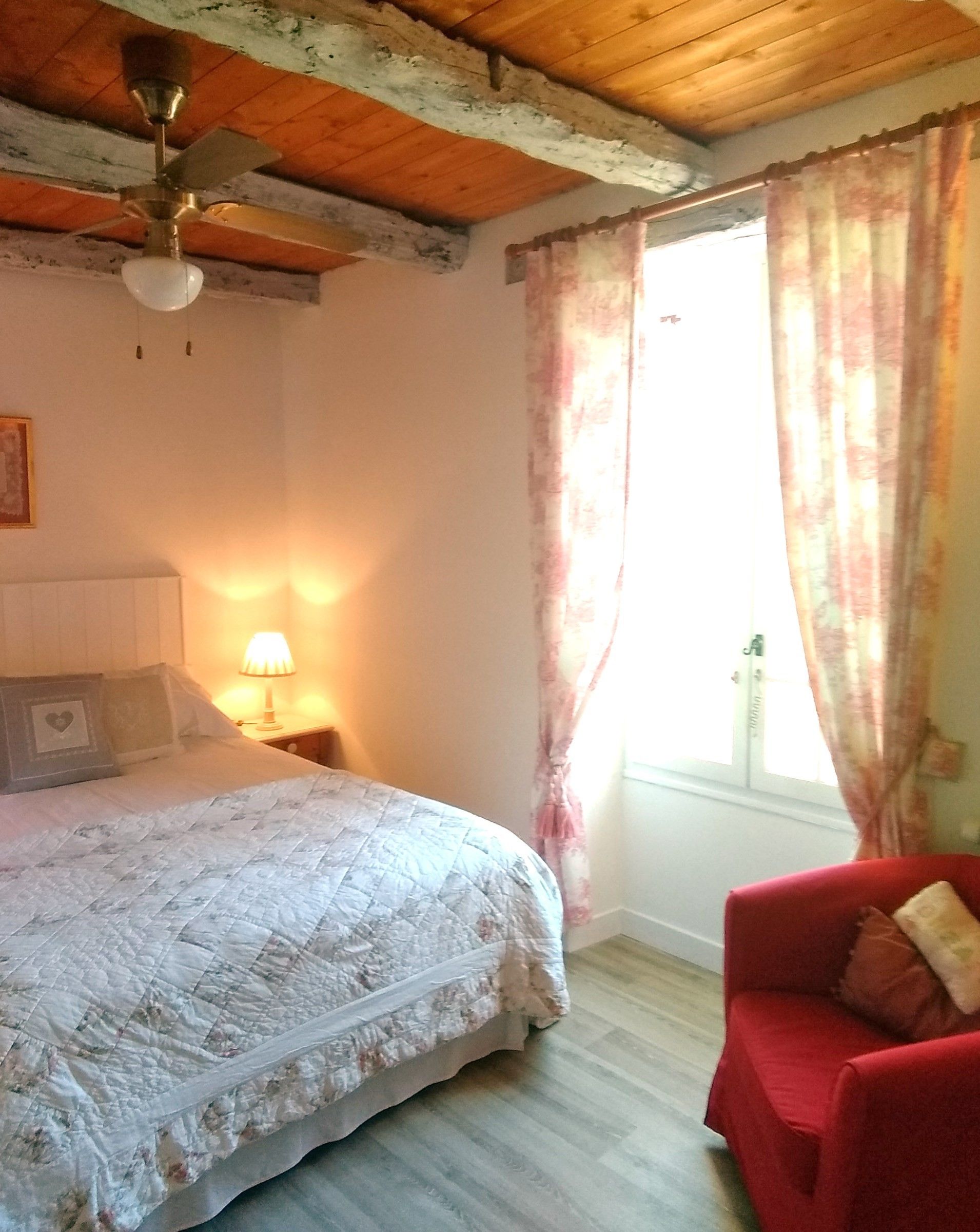 Une chambre à coucher avec un lit, une chaise rouge, un ventilateur de plafond, une fenêtre avec des rideaux et une lampe fixée au mur.