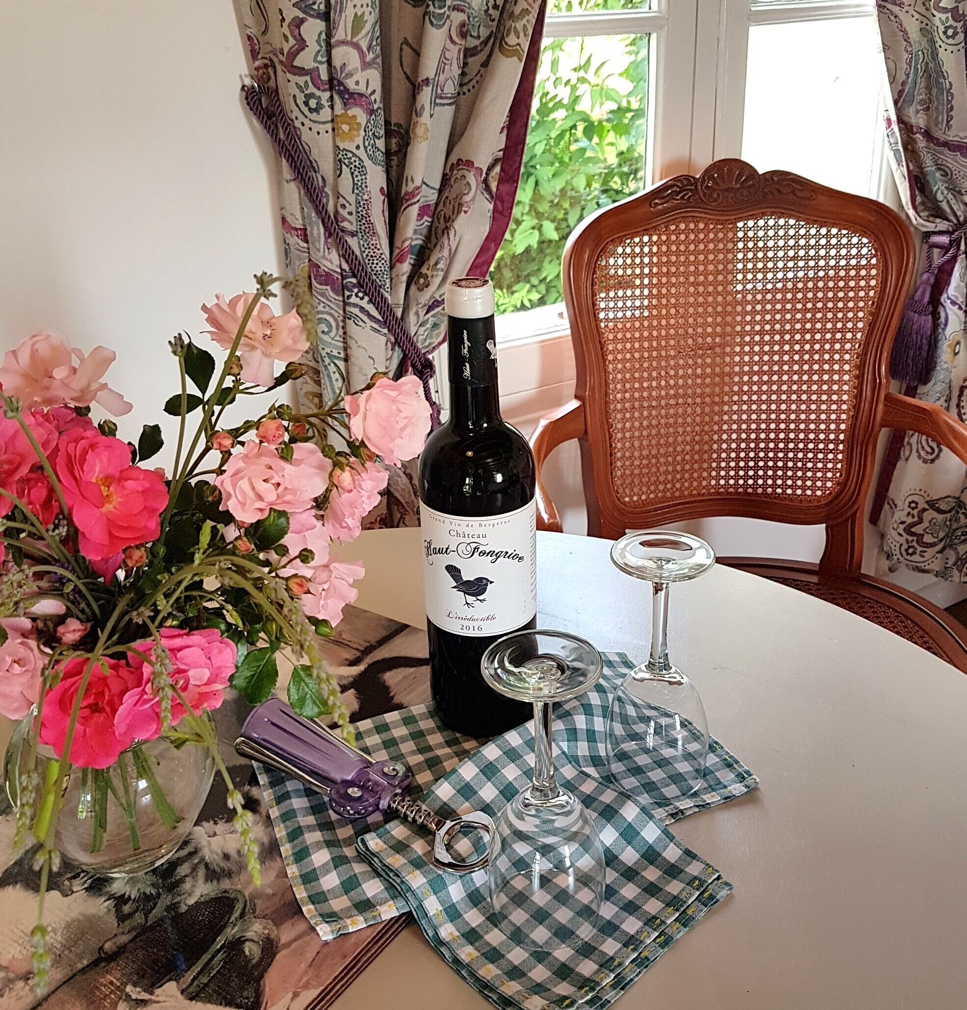 En vinflaske, et glas, en korkskrue og en ternet serviet på et bord med en stol i baggrunden.