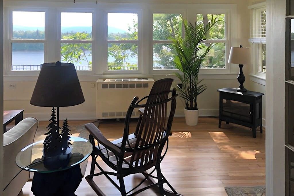 A room with a wooden rocking chair, a small table with a lamp, a potted plant, and a view of a lake through large windows.