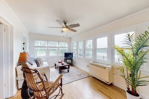 A spacious living room with a ceiling fan, large windows, a television, a fireplace, and a potted plant. - 1BR river view cottage with Hudson River views (Queensbury)