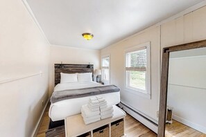 A bedroom with a bed, bedside tables, a bench with towels, and a window with blinds. - 1BR river view cottage with Hudson River views (Queensbury)