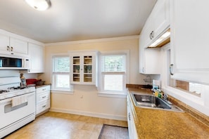A kitchen with white cabinets, a stove, a sink, and two windows with blinds. - 1BR river view cottage with Hudson River views (Queensbury)