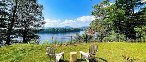 Un espacio con césped, dos sillas y una mesa pequeña, con vista a un lago, árboles y una valla al fondo.