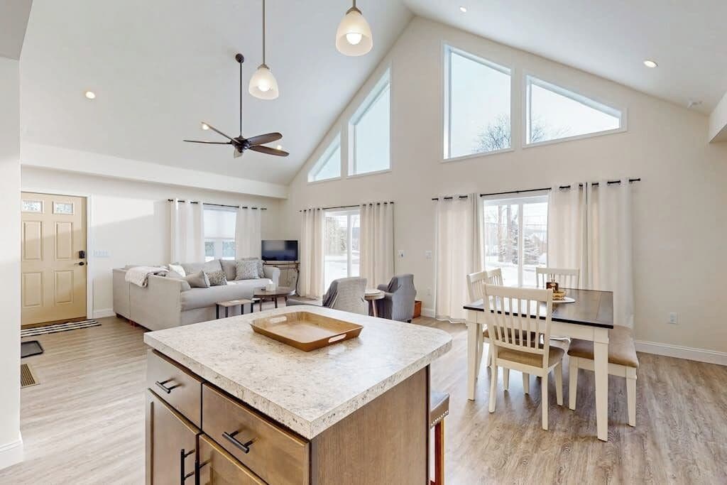 A spacious living area with a high ceiling, a large window, a dining table, and a kitchen island.