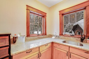 A kitchen with wooden cabinets, granite countertops, a stainless steel sink, and two windows with a view of a snowy landscape. - Luxe 4BR Mountainview Adirondacks (North Creek)