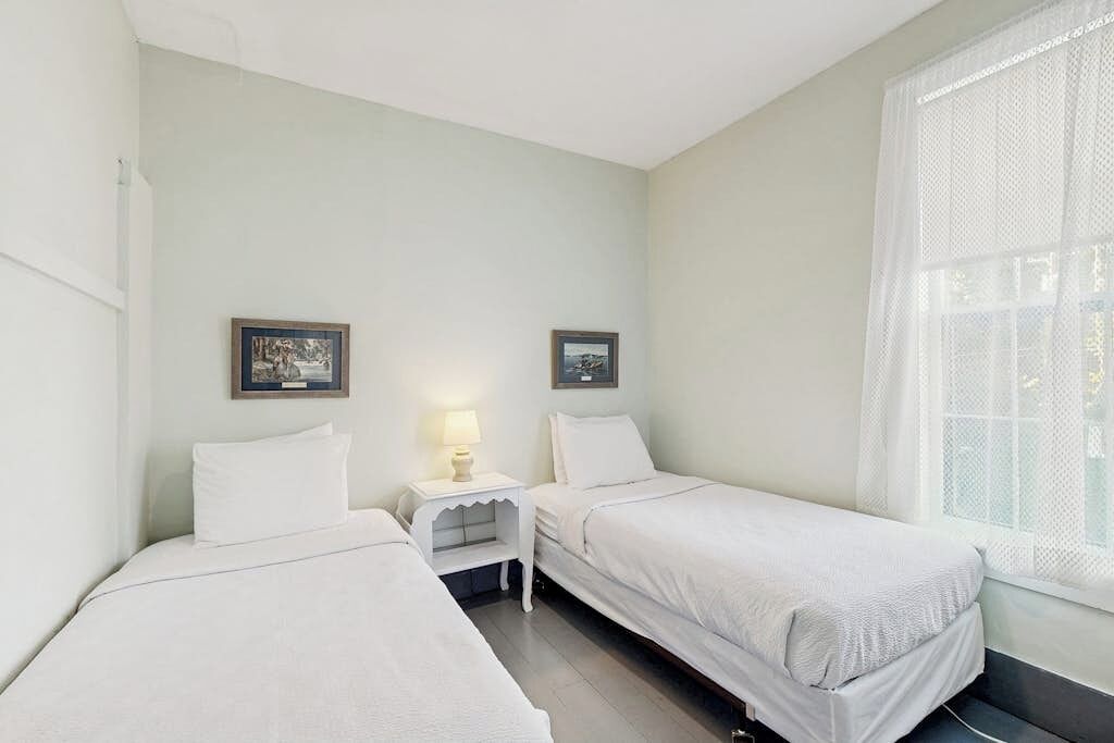 Una habitación con dos camas individuales, una mesita de noche pequeña con lámpara y una ventana con cortinas translúcidas.