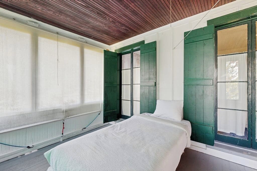 Een kamer met een bed, een raam met groene luiken, een radiator en een houten plafond.