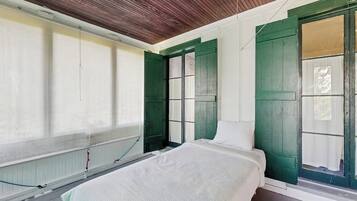 Een kamer met een bed, een raam met groene luiken, een radiator en een houten plafond.