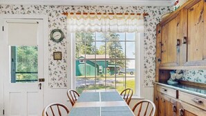 Un comedor con una mesa, sillas, un armario de madera y una ventana con vista a una casa verde.