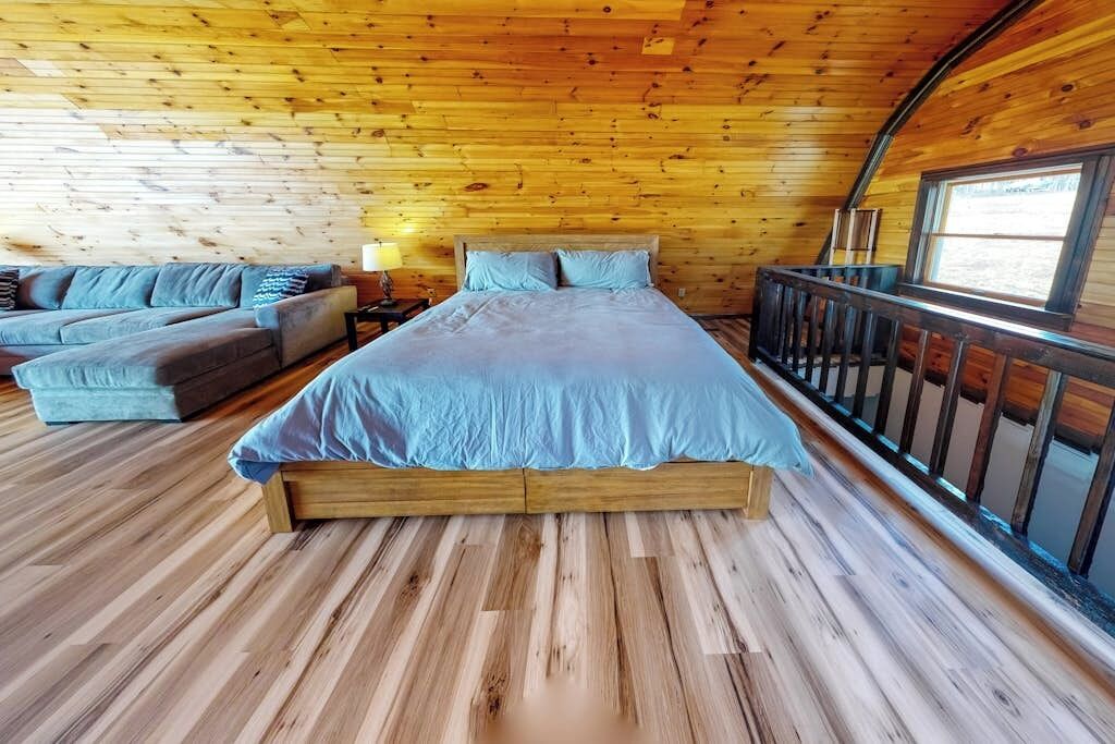 A bedroom with a wooden ceiling, a bed with a blue bedspread, a sofa set, and a window with a view.