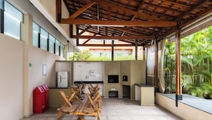 Uma área de refeições ao ar livre com uma longa mesa de madeira e cadeiras, um espaço de cozinha com geladeira e micro-ondas, e vista para o verde através de grandes janelas.
