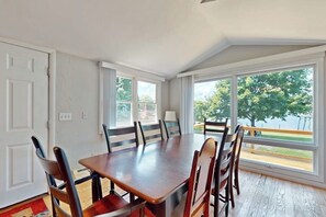 A dining area with a wooden table and chairs, large windows with a view of trees, and a white door.