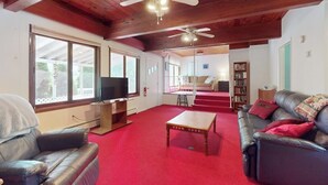 Ein geräumiges Wohnzimmer mit rotem Teppich, Ledersofas, einem Holztisch, Deckenventilatoren und einem Fernseher.