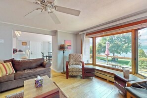 A living room with a ceiling fan, a large window with a view of a deck and trees, a brown leather sofa, and a patterned armchair.