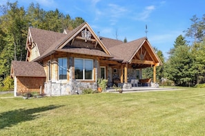 A two-story log cabin with a stone foundation, large windows, and a covered porch. - 3 BR riverfront house with wood stove - near skiing (North Creek)