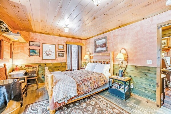 A cozy room with a wooden bed, a small table, and a chair. There is a lamp on the wall and a picture frame. - Dog-friendly 2BR cabin with mountain view (Indian Lake)