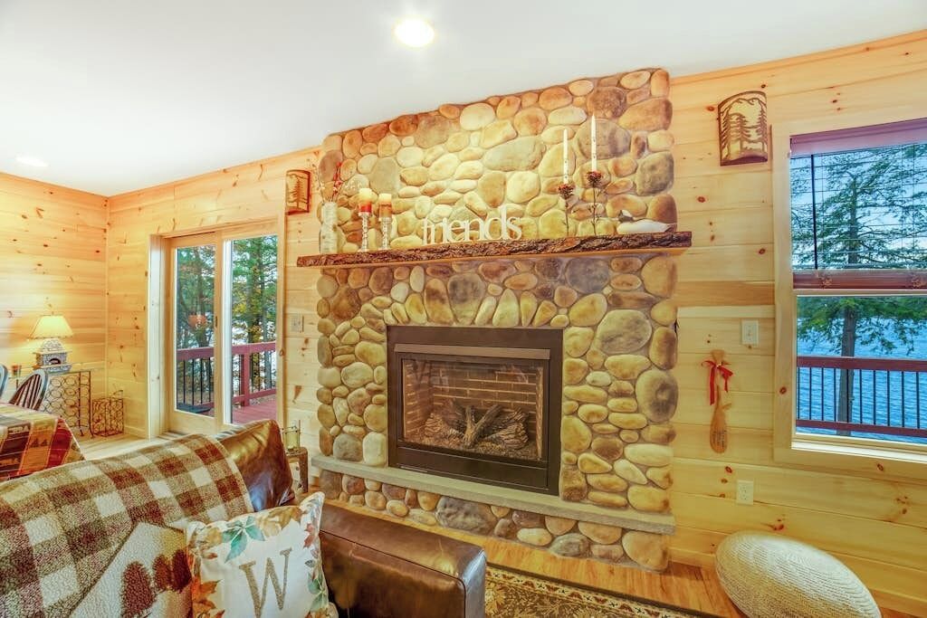 A cozy cabin interior with a stone fireplace, wooden walls, and a view of the outdoors.