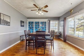 Une salle à manger avec une table en bois, des chaises, un ventilateur de plafond et une vue dégagée sur l’extérieur grâce à de grandes fenêtres.