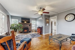 Un salon avec une cheminée, un ventilateur de plafond, une horloge et une grande fenêtre.