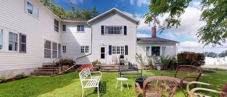 A two-story house with a front porch, a small garden with chairs, and a clear sky.