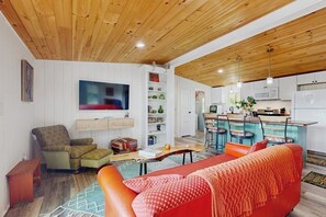 Una sala de estar moderna con sofá, sillón y televisión, contigua a una cocina con taburetes de bar y techo de madera.
