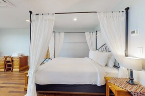 Ein Bett mit Baldachin, ausgestattet mit weißer Bettwäsche und Vorhängen, ein Nachttisch mit Lampe sowie ein Holznachtkästchen.