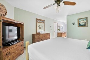 A bedroom with a bed, wooden dresser, television, ceiling fan, and a bathroom visible through an open door.