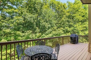 A wooden deck with a round table and chairs, surrounded by a railing, overlooking a dense green forest.
