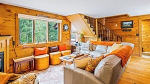 L’intérieur d’un chalet accueillant, avec une cheminée, des murs en bois, un escalier et une grande fenêtre donnant sur un paysage verdoyant.