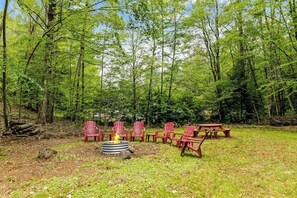 A forested campsite with a fire pit, red chairs, and picnic tables.