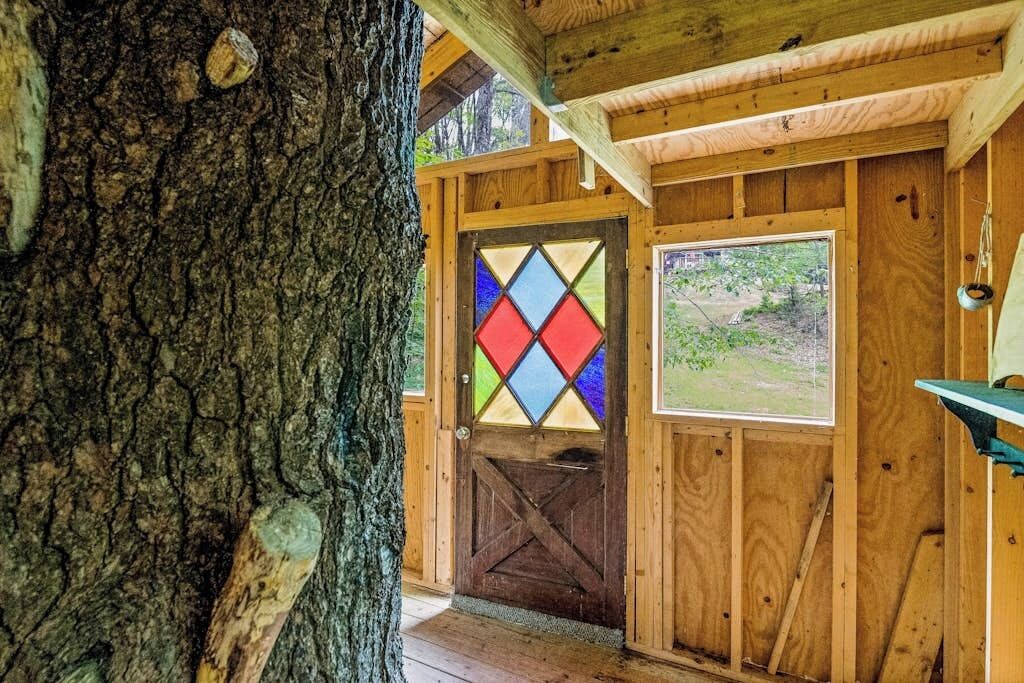 A wooden cabin with a large tree trunk on the left, a colorful diamond-patterned door, and a window with a view of the outdoors.