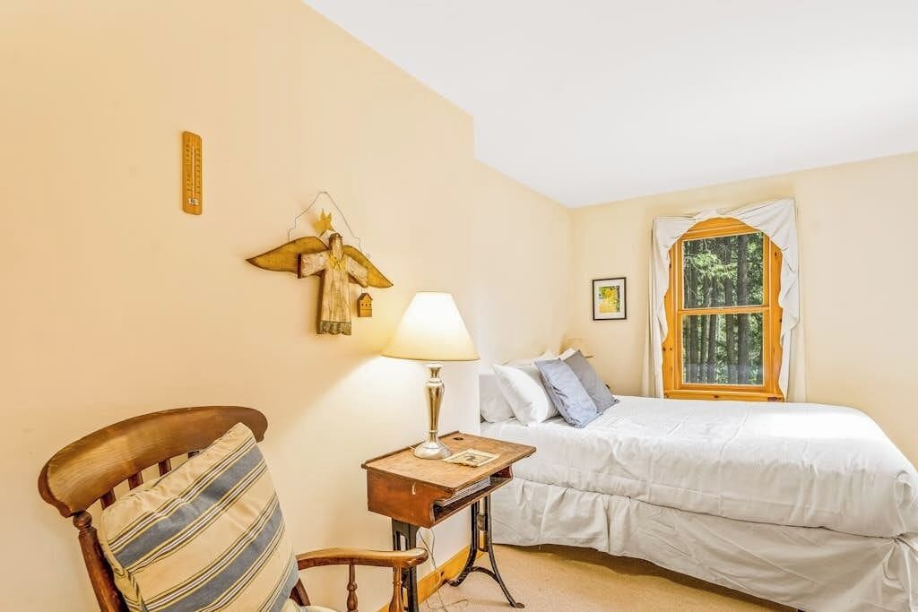 Ein Schlafzimmer mit einem Bett, einem Sessel, einem Nachttisch, einer Lampe, einem Fenster und einer Wanddekoration.