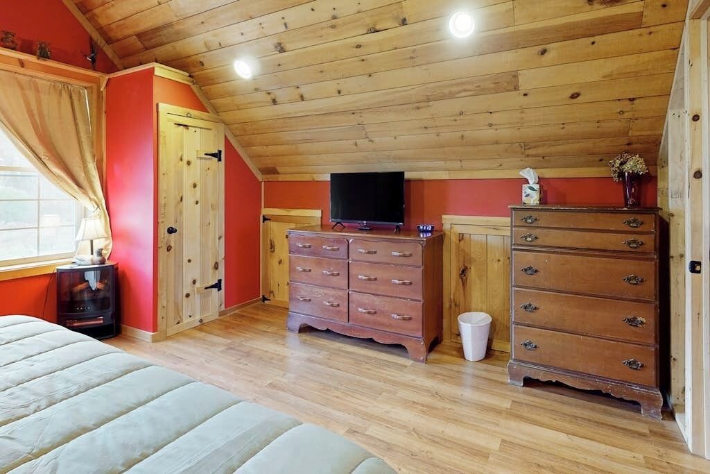 Una habitación con techo de madera, cama, cómoda, televisor y ventana con cortinas.