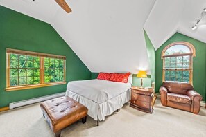 A bedroom with a sloped ceiling, a large bed, a nightstand, a leather armchair, and a window with a view of trees. - Quiet 4BR Adirondacks Dog Friendly - Hot Tub (Johnsburg)